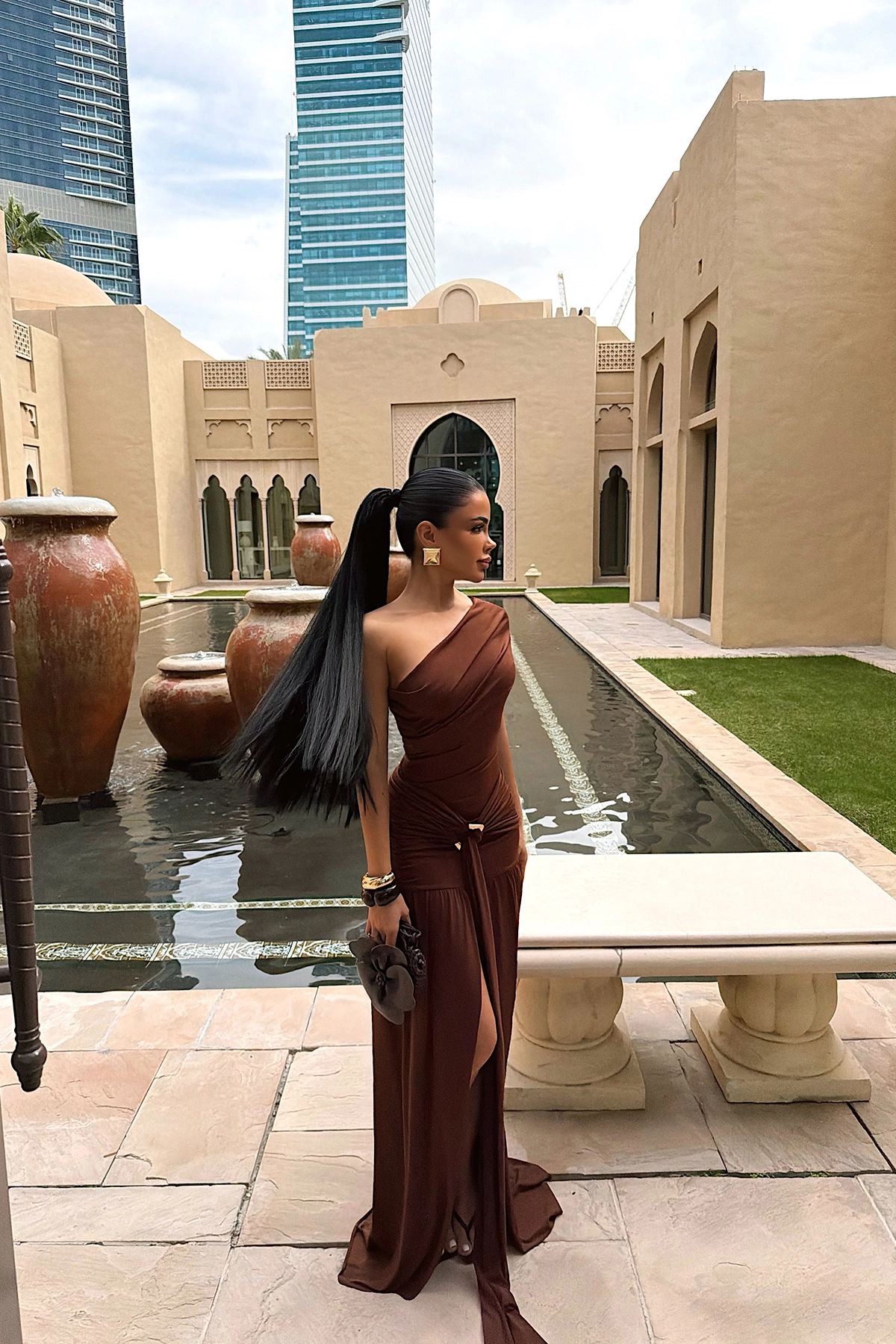 brown maxi dress featuring an elegant one-shoulder neckline, flattering ruched detailing and a softly draped skirt. Its refined silhouette creates an effortlessly glamorous look, perfect for evening events, weddings and special occasions.